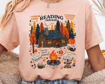 Op de afbeelding: Een perzikkleurig T-shirt met een kleurrijke illustratie van een gezellige hut in een bosrijke omgeving. Het ontwerp bevat een kampvuur, boeken en de woorden "Reading Retreat" en "For the Cozy Readers". Het shirt wordt gecombineerd met een spijkerbroek.
