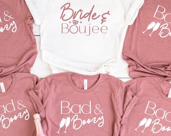 Op de afbeelding: Zes roze T-shirts met witte tekst. Eén shirt zegt "Bride & Boujee" en de andere vijf shirts zeggen "Bad & Boozy" met een afbeelding van twee champagne glazen.
