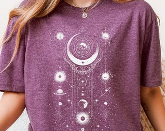 May include: A purple t-shirt featuring a white celestial design with a crescent moon, planets, and stars. The shirt is made of a soft material and is a comfortable fit. The design is a detailed, intricate graphic.