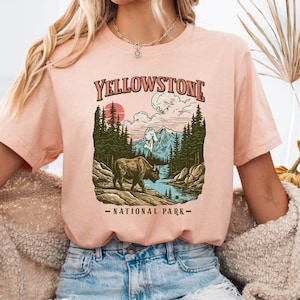 Puede incluir: Una camiseta color melocotón con un gráfico del Parque Nacional de Yellowstone. El diseño incluye un bisonte, montañas, árboles y el texto "YELLOWSTONE NATIONAL PARK". La camiseta es de manga corta y la lleva una persona.