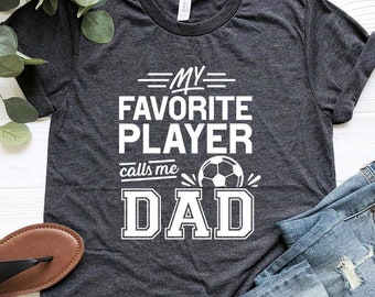 Puede incluir: Una camiseta gris oscuro con texto blanco que dice "Mi jugador favorito me llama papá" con un gráfico de balón de fútbol. La camiseta está colocada sobre una superficie de madera blanca con un par de sandalias marrones y unos vaqueros azules.