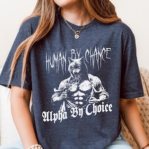 Puede incluir: Camiseta azul oscuro con un gráfico blanco de una figura híbrida lobo-humano. El texto "HUMAN BY CHANCE" está encima de la figura, y "Alpha By Choice" debajo. La camiseta tiene mangas cortas y cuello redondo.
