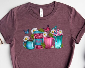 Puede incluir: Una camiseta de color marrón rojizo con un gráfico de una taza de café, una taza y un vaso con una pajita. La taza de café tiene la palabra "Mom" en ella. Hay mariposas y flores alrededor de las tazas.