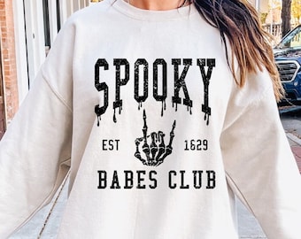 May include: A white sweatshirt with a black graphic print that says "Spooky Babes Club" with a skeleton hand making a rock and roll sign. The text also says "Est 1629".