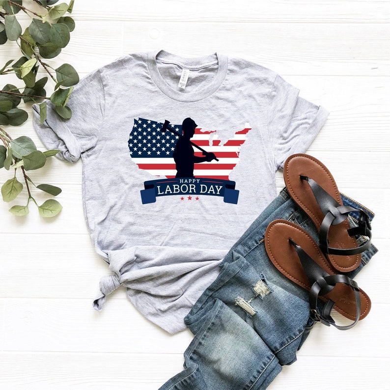 May include: A gray t-shirt with a graphic of a silhouette of a person holding an axe in front of an American flag in the shape of the United States. The text "Happy Labor Day" is on a blue ribbon below the graphic.