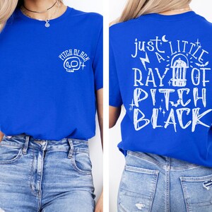 May include: Royal blue t-shirt with white graphic text and a skull design. The front of the shirt has the words "Pitch Black" with a skull graphic. The back of the shirt has the phrase "Just a Little Ray of Pitch Black".