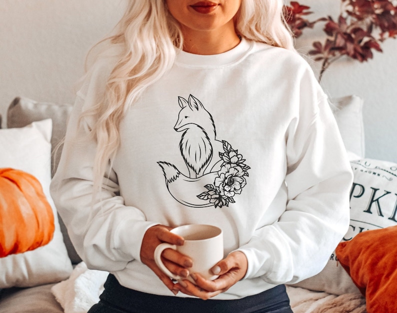May include: White sweatshirt with a black line drawing of a fox curled up with flowers. The fox is facing left and has a bushy tail.