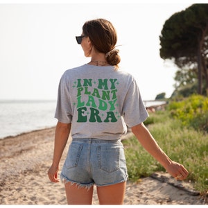 May include: A gray t-shirt with the text "IN MY PLANT LADY ERA" in green and white letters with stars on either side of the text.