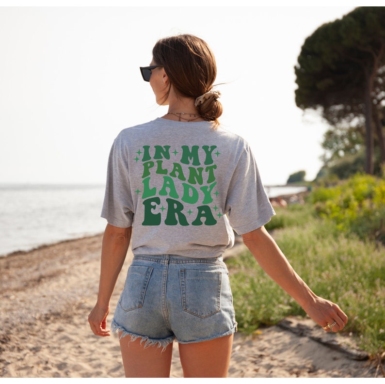 May include: A gray t-shirt with the text "IN MY PLANT LADY ERA" in green and white letters with stars on either side of the text.