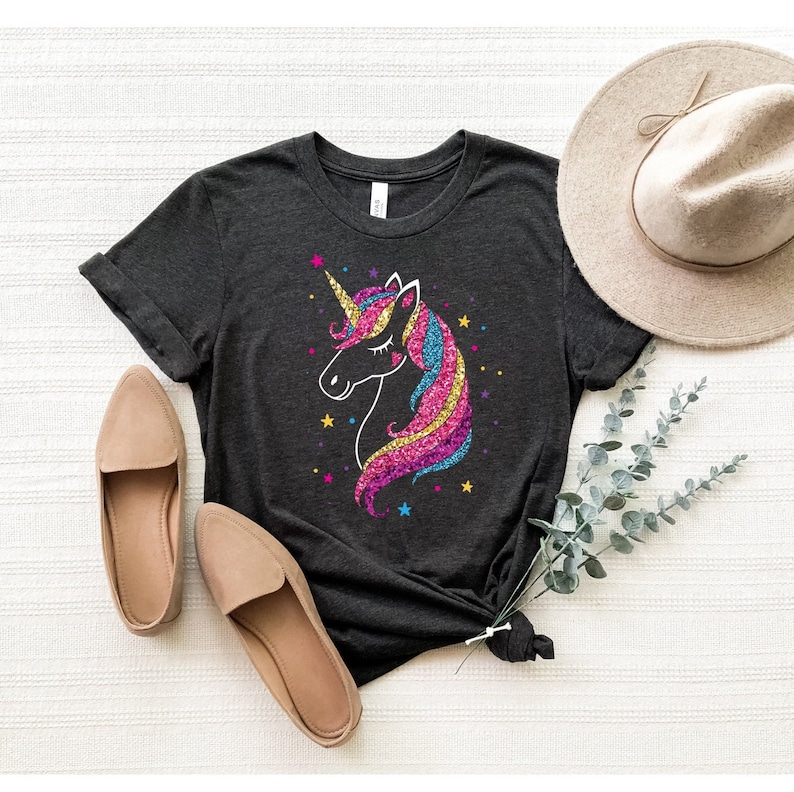 以下が含まれることがあります： キラキラ光るユニコーンのグラフィックがプリントされたダークグレーのTシャツ。ユニコーンはピンク、ブルー、イエローのたてがみと尻尾をしています。グラフィックは小さな星で囲まれています。