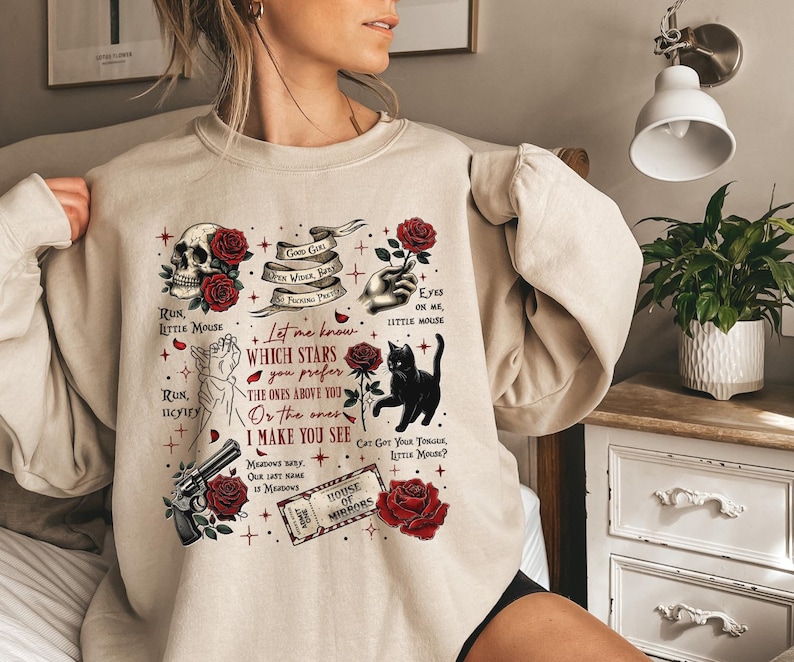 May include: Beige sweatshirt with a graphic design featuring a skull, roses, a black cat, a revolver, and text. The design includes phrases like "Run, Little Mouse" and "House of Mirrors" in a gothic style.