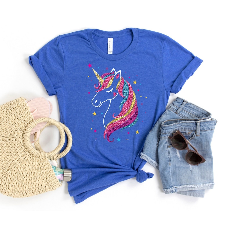 以下が含まれることがあります： ロイヤルブルーのTシャツに、キラキラ光るユニコーンのデザインが施されています。ユニコーンはピンク、イエロー、ブルーのたてがみと白い角をしています。Tシャツはデニムのショートパンツと麦わら帽子と合わせています。