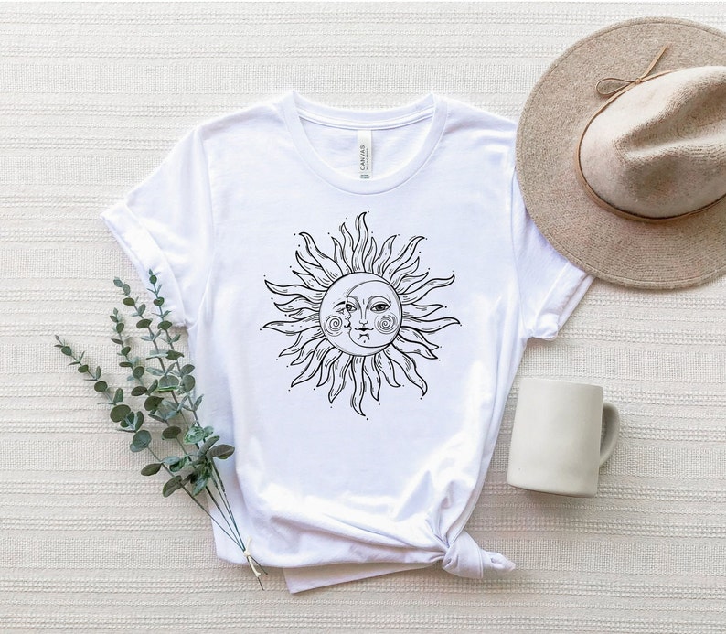 K&ouml;nnte beinhalten: Wei&szlig;es T-Shirt mit einem schwarzen und wei&szlig;en Sonnen- und Mondmotiv. Sonne und Mond sind stilisiert mit Gesichtern und haben eine sichelf&ouml;rmige Mondform.