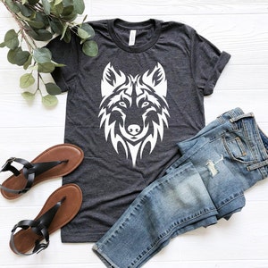 May include: A dark gray t-shirt with a white wolf graphic design. The wolf is stylized with a tribal design. The t-shirt is laying on a white surface with a pair of brown sandals and blue jeans.
