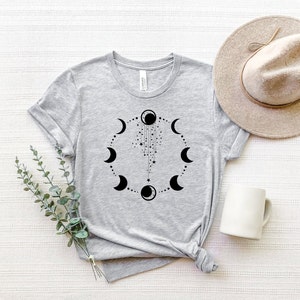Puede incluir: Una camiseta gris con un dise&ntilde;o negro de fases lunares. El dise&ntilde;o presenta un c&iacute;rculo de lunas en diferentes fases, con una cadena de estrellas y una luna creciente colgando de la luna llena.
