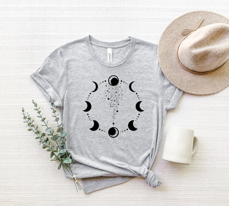Puede incluir: Una camiseta gris con un dise&ntilde;o negro de fases lunares. El dise&ntilde;o presenta un c&iacute;rculo de lunas en diferentes fases, con una cadena de estrellas y una luna creciente colgando de la luna llena.