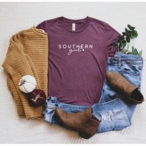 Op de afbeelding: Een maroon t-shirt met de tekst "SOUTHERN girl" in witte letters. Het shirt ligt op een wit oppervlak met een bruine trui, een blauwe jeans en bruine laarzen.