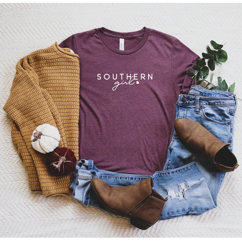 Op de afbeelding: Een maroon t-shirt met de tekst "SOUTHERN girl" in witte letters. Het shirt ligt op een wit oppervlak met een bruine trui, een blauwe jeans en bruine laarzen.