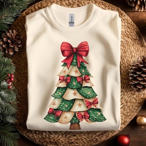 Puede incluir: Sudadera de color crema con un diseño de árbol de Navidad en acuarela. El árbol está compuesto por capas verdes y crema, adornado con lazos rojos y estrellas doradas. Un gran lazo rojo corona el árbol. La sudadera se muestra sobre una superficie tejida.