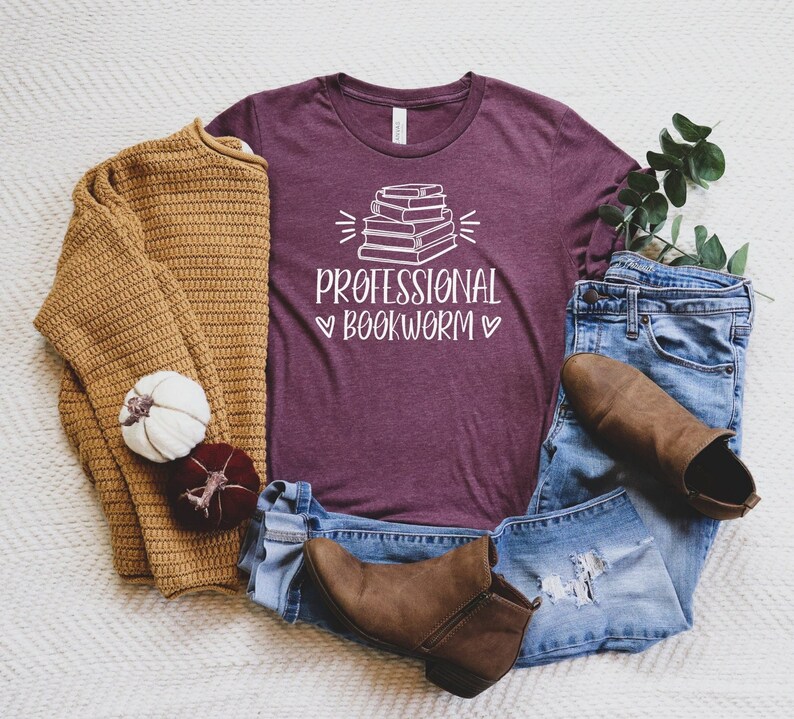 Peut inclure: Un t-shirt marron avec une pile de livres et le texte "Professional Bookworm" en blanc. Le t-shirt est pos&eacute; sur une surface blanche avec un pull marron, un jean bleu et des bottes marron.