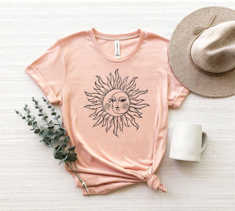 K&ouml;nnte beinhalten: Ein pfirsichfarbenes T-Shirt mit einem schwarzen und wei&szlig;en Sonnen- und Mondmotiv. Die Sonne hat ein Gesicht mit geschlossenen Augen und einen Halbmond als Nase.
