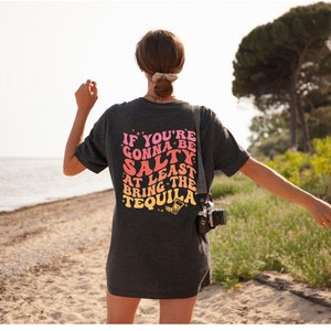 May include: Dark gray t-shirt with the text "IF YOU'RE GONNA BE SALTY AT LEAST BRING THE TEQUILA" in pink, orange, and yellow. The shirt is worn by a person on a beach, with a camera slung over their shoulder.