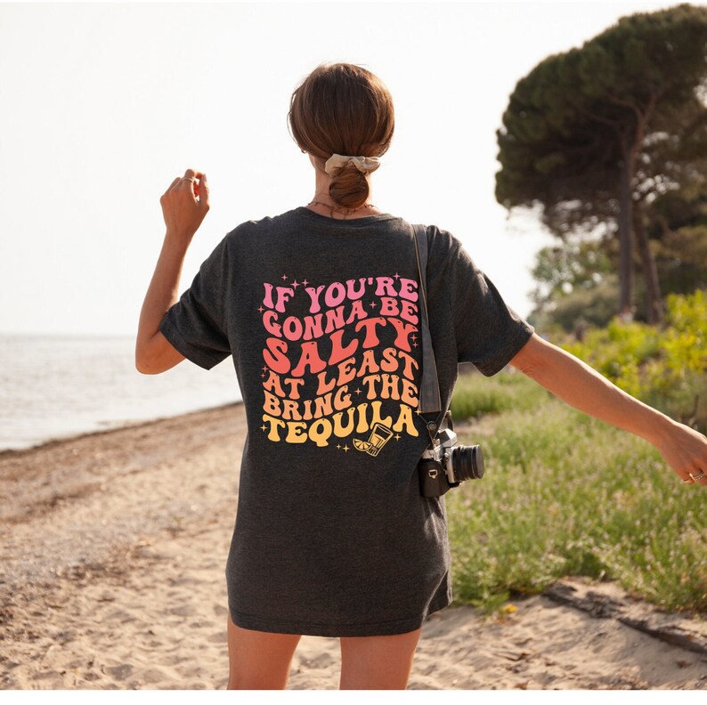 May include: Dark gray t-shirt with the text "IF YOU'RE GONNA BE SALTY AT LEAST BRING THE TEQUILA" in pink, orange, and yellow. The shirt is worn by a person on a beach, with a camera slung over their shoulder.