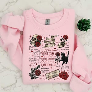 May include: A light pink sweatshirt with a graphic design featuring a skull, roses, a black cat, a gun, and text. The text includes phrases like "Run, Little Mouse" and "House of Mirrors."