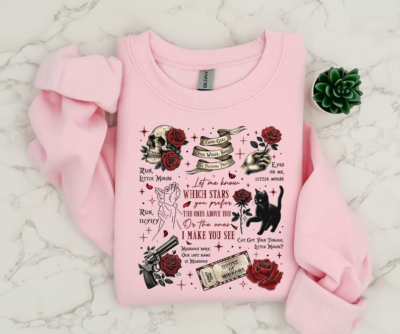 May include: A light pink sweatshirt with a graphic design featuring a skull, roses, a black cat, a gun, and text. The text includes phrases like "Run, Little Mouse" and "House of Mirrors."