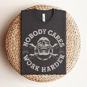 Puede incluir: Camiseta gris oscuro con cuello redondo, con un gráfico desgastado. El gráfico incluye una calavera, llaves inglesas y el texto "NOBODY CARES WORK HARDER" en un diseño circular. La camiseta se muestra sobre una superficie tejida.