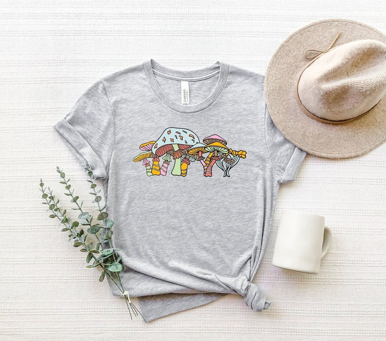 May include: A gray t-shirt with a colorful mushroom graphic. The mushrooms are drawn in a whimsical style with orange, pink, blue, and green accents.