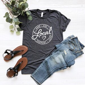 May include: A dark gray t-shirt with a white circle graphic that says "Shop Eat Local Support Love" with a heart in the center. The shirt is laid flat on a white wooden surface with a pair of black sandals and blue jeans.
