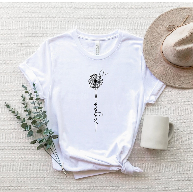 May include: White t-shirt featuring a black dandelion graphic with the word "vegan" written vertically beneath it.