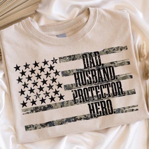 May include: A beige t-shirt featuring a distressed American flag design. The flag's stripes are camouflage, and the text reads "DAD. HUSBAND. PROTECTOR. HERO." Black stars represent the stars on the flag.