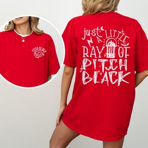 May include: Red t-shirt with white text and graphics. The front left chest has a skull graphic with the words "Pitch Black". The back of the shirt has the phrase "Just a little ray of Pitch Black" with a graphic of a vintage radio.