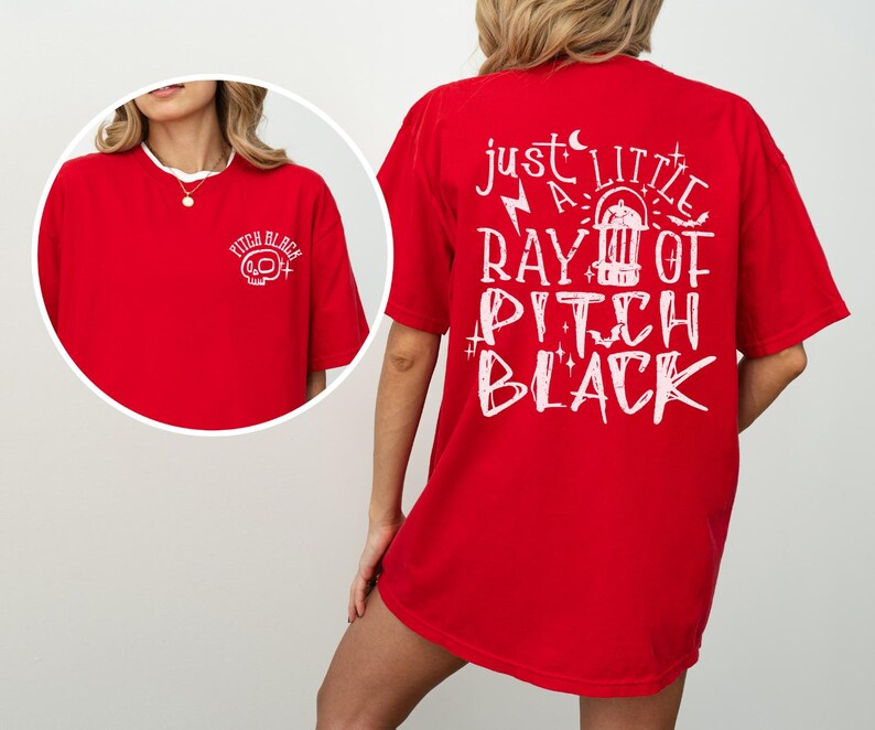 May include: Red t-shirt with white text and graphics. The front left chest has a skull graphic with the words "Pitch Black". The back of the shirt has the phrase "Just a little ray of Pitch Black" with a graphic of a vintage radio.