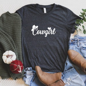 K&ouml;nnte beinhalten: Ein dunkelgraues T-Shirt mit einer wei&szlig;en Grafik eines Cowboyhuts und dem Wort "Cowgirl". Das Shirt liegt auf einer wei&szlig;en Oberfl&auml;che mit einem gr&uuml;nen Pullover, einer blauen Jeans und braunen Stiefeln.