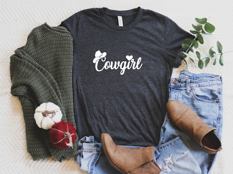 K&ouml;nnte beinhalten: Ein dunkelgraues T-Shirt mit einer wei&szlig;en Grafik eines Cowboyhuts und dem Wort "Cowgirl". Das Shirt liegt auf einer wei&szlig;en Oberfl&auml;che mit einem gr&uuml;nen Pullover, einer blauen Jeans und braunen Stiefeln.