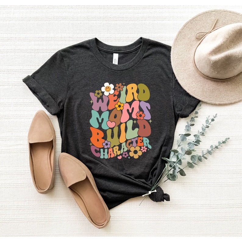 May include: A dark gray t-shirt with a colorful retro-style graphic that says "Weird Moms Build Character". The graphic features flowers and a heart.