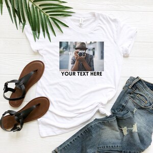 May include: White t-shirt with a photo of a person wearing a hat and holding a camera. The text "YOUR TEXT HERE" is printed below the photo.