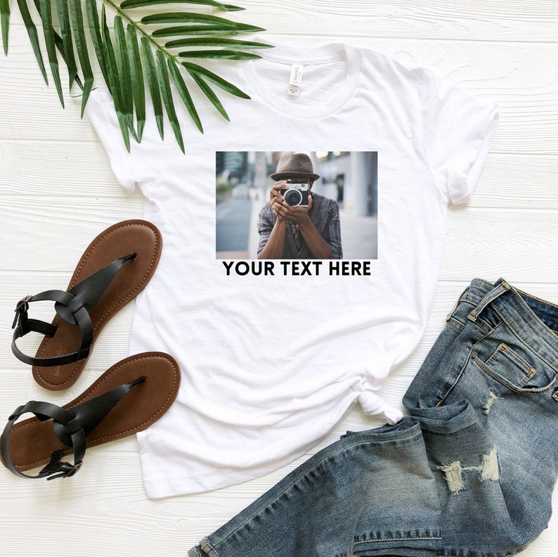 May include: White t-shirt with a photo of a person wearing a hat and holding a camera. The text "YOUR TEXT HERE" is printed below the photo.