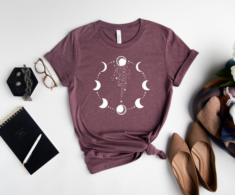 Puede incluir: Una camiseta de color marr&oacute;n rojizo con un gr&aacute;fico blanco de un c&iacute;rculo de lunas y estrellas. El gr&aacute;fico est&aacute; centrado en la camiseta.
