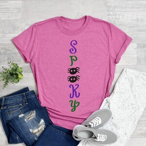 May include: A pink t-shirt with the word "Spooky" spelled vertically down the front. The letters are in different colors, with the "S" in purple, the "P" in green, the "O" in black, the "K" in purple, and the "Y" in green. There are two black spiders between the "O" and the "K".
