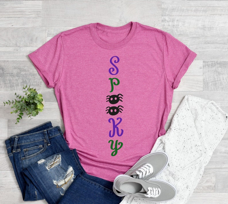 May include: A pink t-shirt with the word "Spooky" spelled vertically down the front. The letters are in different colors, with the "S" in purple, the "P" in green, the "O" in black, the "K" in purple, and the "Y" in green. There are two black spiders between the "O" and the "K".