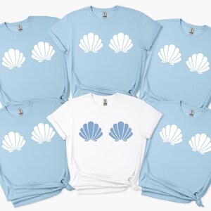Könnte beinhalten: Eine Auswahl an hellblauen und weißen T-Shirts mit Muschelmuster. Ein weißes T-Shirt hat ein blaues Muscheldesign. Die kurzärmeligen Shirts haben einen lässigen Stil, passend für Strand oder Sommer.