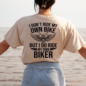 May include: A beige t-shirt with a black graphic that reads "I don't ride my own bike but I do ride my own biker" with a motorcycle and wings design.
