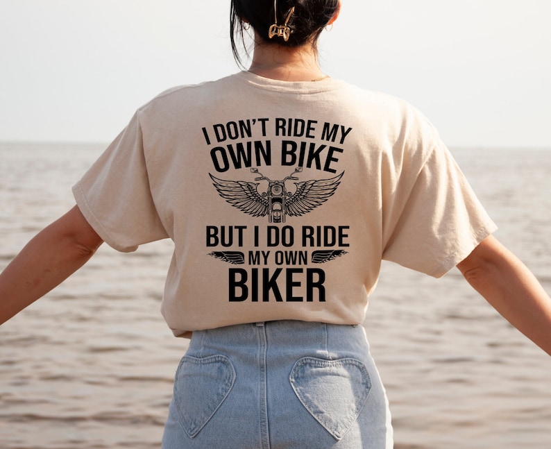 May include: A beige t-shirt with a black graphic that reads "I don't ride my own bike but I do ride my own biker" with a motorcycle and wings design.