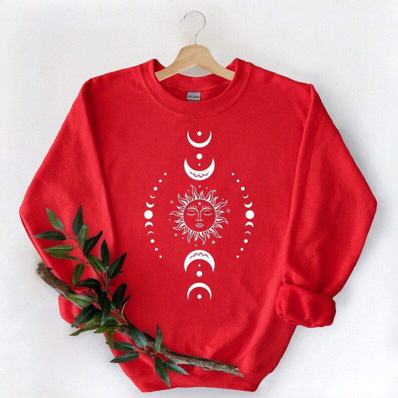 Puede incluir: Sudadera roja con un dise&ntilde;o blanco de sol y luna. El sol tiene una cara y est&aacute; rodeado por un c&iacute;rculo de lunas.