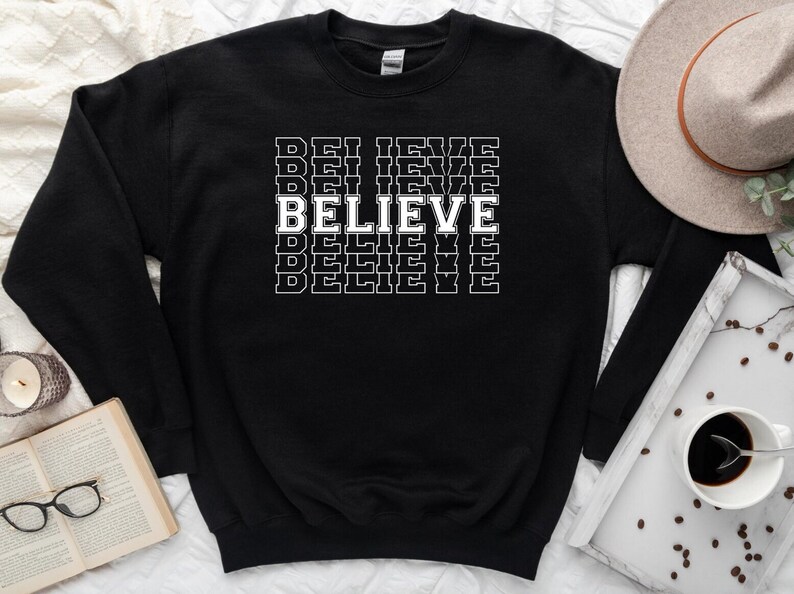 Puede incluir: Sudadera negra con la palabra "BELIEVE" impresa en letras blancas, repetida varias veces en un patr&oacute;n apilado.