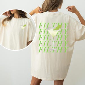 May include: Cream-colored t-shirt featuring a lime green martini glass graphic on the front and the word "FILTHY" repeated in lime green on the back. The shirt has a relaxed fit and short sleeves.